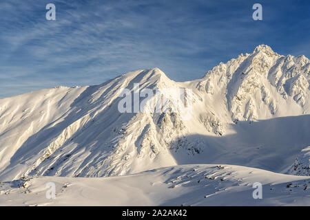 Scenic austrian alpine mountain peaks covered with snow layer in winter at warm sunset or sunrise time. Blue clear sy on background. Natural rocky Stock Photo