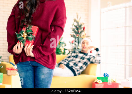 A beautiful Asian woman is surprising his boyfriend by hiding a gift box in the back and walking towards him while the man is lying on the sofa in the Stock Photo