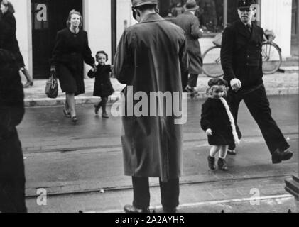 A police officer of the Wilhelmshaven Police stands on a street with his back to the viewer. Pedestrians with children are crossing the road. Stock Photo