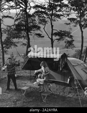 A young couple went camping in the woods with their VW Beetle. While he fastens the tent, she takes care of the folding table and the food. Stock Photo