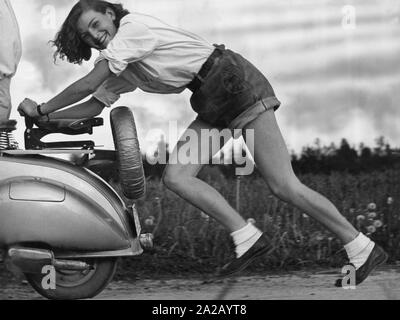 A girl in short leather pants laughs while pushing a scooter. Stock Photo