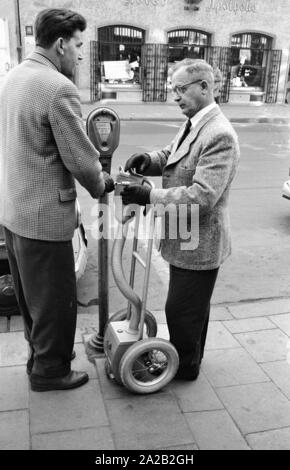 Two officials emptying parking meters installed in the city center of Munich. After opening the parking meter, the coins fall into the small, mobile safe through a kind of plastic hose. The entire construction is reminiscent of a sack truck. Parking meters had been increasingly set up in Munich to ban long-term parkers from the city center since the mid 1950s. Here, the officials connecting the coin tube. Stock Photo