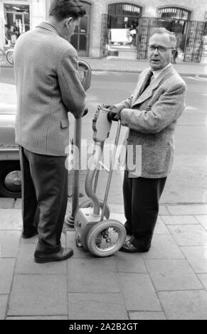 Two officials emptying parking meters installed in the city center of Munich. After opening the parking meter the coins fall into the small, mobile safe through a kind of plastic hose. The entire construction is reminiscent of a sack truck. Parking meters had been increasingly set up in Munich to ban long-term parkers from the city center since the mid 1950s. Photo of officials closing a parking meter after having emptied it. Stock Photo