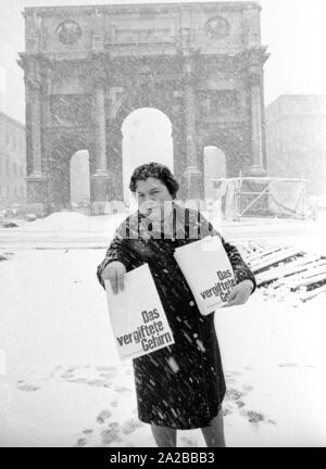 As part of a campaign against narcotics in Munich, Ms. Baumann distributes the publication of the Süddeutsche Zeitung 'Das vergiftete Gehirn' ('The Poisoned Brain') to passers-by. She stands in dense snow on the Leopoldstraße, behind her is the Siegestor. Stock Photo