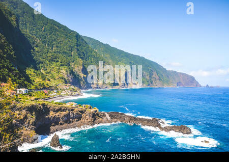 Stunning cliffs on the northern coast of Madeira Island, Portugal. Steep green rocks, small city and dark blue sea water photographed by village Seixal. Tourist destinations. Atlantic ocean. Stock Photo