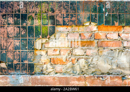 Vintage weathered orange brick wall with remains of cement and plaster, broken layer of old glossy black ceramic vertical tile with spots of red-pink Stock Photo