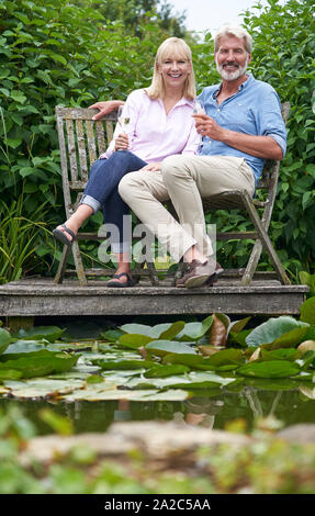 Portrait Of Mature Couple Celebrating With Champagne Sitting On Chairs On Wooden Jetty By Lake Stock Photo
