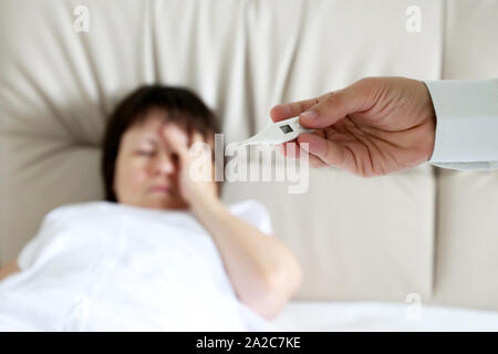 Fever and illness, sick woman lying in a bed, digital thermometer in male hand close up. Doctor measuring body temperature of patient, cold anв flu Stock Photo