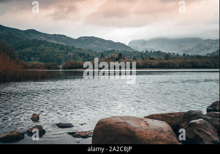 Looking out across a calm lake shoreline towards large mountains at sunrise Stock Photo