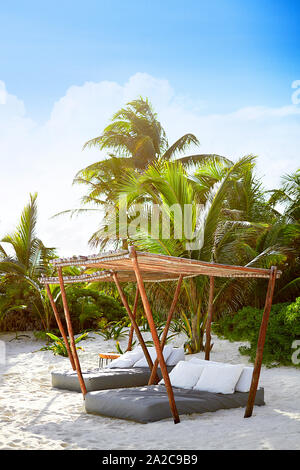 Lounge beds shaded under canopies on white sandy beach with palm trees in background Stock Photo