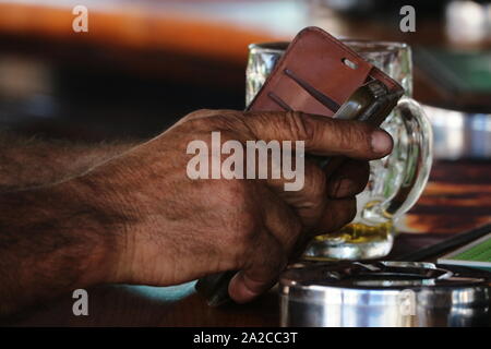 Close up of working man's hands using smart phone in leather case with partially drunk glass of beer and stainless steel ashtray on wood bar top Stock Photo