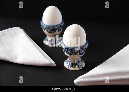 Still life of two white eggs in egg stands with white napkins near it on day sunlight. International world egg day concept on dark background Stock Photo