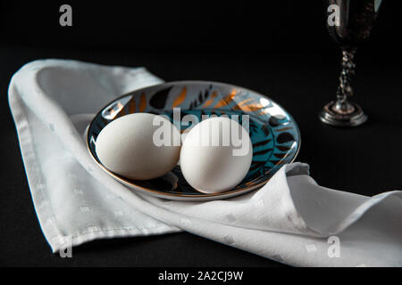 Still life of beautiful saucer with two eggs on white napkin and sterling silver wine glass near on dark background. International world egg day. Stock Photo