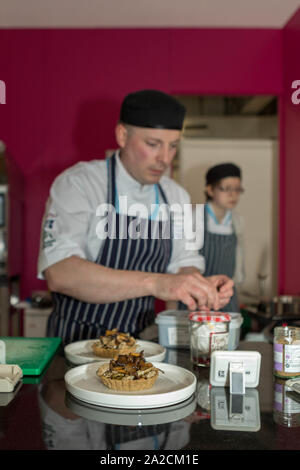 Olympia, London, UK. 2nd Oct, 2019. A chef in white coat, black hat, and striped apron prepares a fish on a cutting board in a vibrant professional kitchen. Wearing blue gloves, the chef focuses on cleaning the fish, while another chef works in the background. The kitchen features brightly coloured walls in green and pink, adding visual energy to the culinary workspace. The restaurant trade show at Olympia, Kensington. The event has over 450 exhibitors owning, operating or working in restaurants, hotels, pubs, bars and hospitality establishments across the UK. Penelope Barritt/Ģ����ý Live News Stock Photo