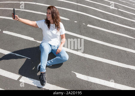 Young woman makes a selfie sitting on the grey floor with white lines. Stock Photo