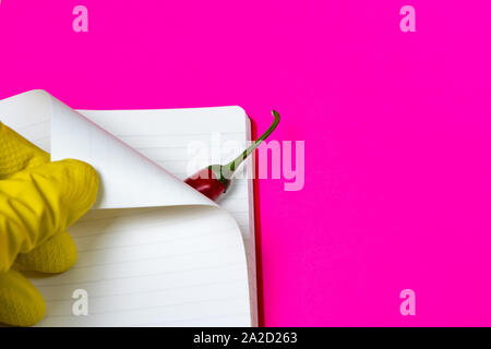 Hands in rubber gloves carefully turn over the page of an empty magazine and open burning red pepper. Bright pink and yellow background. Stock Photo