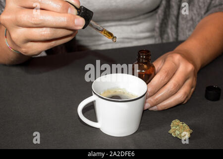 Pouring CBD tincture in a coffee cup, natural medicine made with marihuana. Woman using cannabis oil with a dropper in a table with marihuana bud. Stock Photo