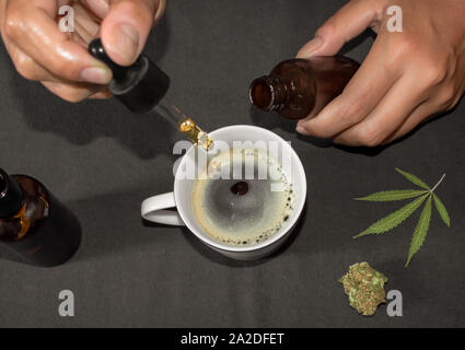 Pouring CBD tincture in a coffee cup, natural remedy of marihuana. Person using cannabis oil with a dropper in a table with marihuana leaf. Stock Photo