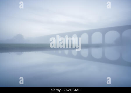 The outline of Arthington Viaduct is visible in thick late autumn fog, reflected in the calmly flowing waters of the River Wharfe before sunrise. Stock Photo
