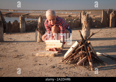 Mature middle aged blonde woman collects firewood for a bonfire on the beach, selective focus Stock Photo