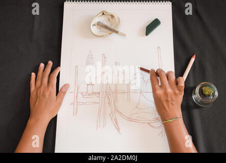 First person view of a woman drawing with a pencil while smoking a marijuana joint. Black table with sketch, casual shell ashtray and cannabis. Stock Photo