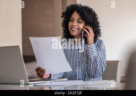 Smiling female entrepreneur talking on her cellphone and reading documents Stock Photo