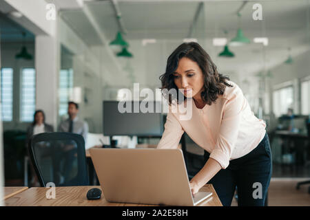 Businesswoman leaning over her office desk working on a laptop Stock Photo