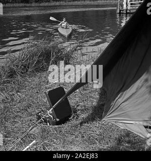 Ein Mann paddelt in einem Klepper Faltboot auf das Ufer zu, wo ein Klepper Zelt aufgebaut ist, Deutschland 1930er Jahre. A man paddling in his Klepper folding boat to the shore of a lake, where a Klepper tent is built, Germany 1930s. Stock Photo