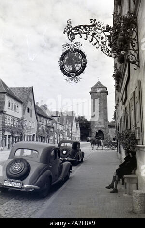 Zwei Schuljungen, die auf einer Bank vor zwei Autos sitzen, schauen einem älteren Verkehsmittel, der Pferdekutsche hinterher, Rothenburg ob der Tauber, Deutschland 1930er Jahre. Two schoolboys sitting on a bench in front of two cars, are watching a much older vehicle passing by, a horse-drawn carriage, Rothenburg ob der Tauber, Germany 1930s. Stock Photo