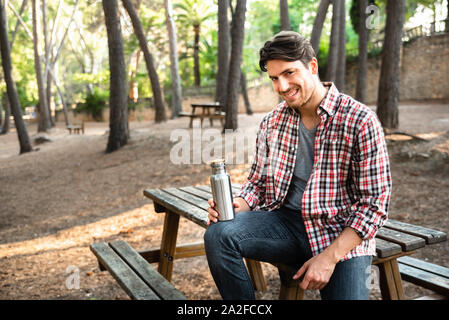 Model man holds a reusable aluminum bottle for water sitting in a park with trees. Stock Photo