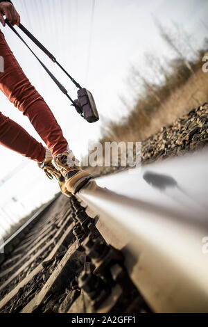 Young woman with legs, dressed in boots walking on railroad tracks, holding a camera in her hands