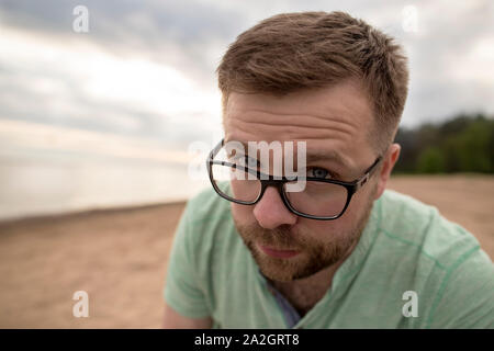 Bearded man in glasses looks questioningly blue eyes frowning, on the sandy sea beach on a cloudy evening. Stock Photo