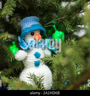 A cute knitted homemade snowman in a hat and scarf sits on the branches of a Christmas tree, which is decorated with a multi-colored garland. Stock Photo