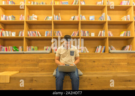 Asian male student using laptop at wooden bookshelf in library  Stock Photo