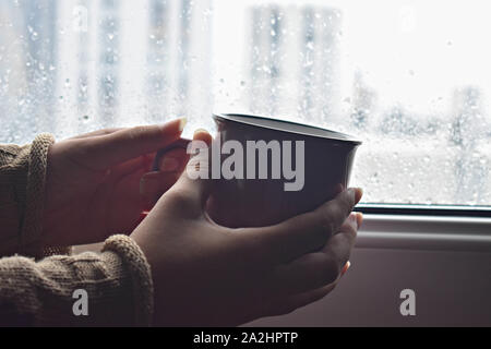 Hand holding hot cup of coffee or tea in morning sunlight on a background of wet window with raindrops / comfortable coffee break Stock Photo
