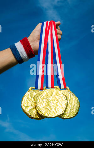 Hand of American athlete holding gold medals hanging from USA colors red, white, and blue ribbon against a bright blue sky background Stock Photo