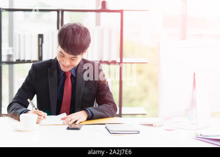 A young Asian businessman wears a suit is sitting signing documents paper with a pen on a desk in the office. He smiled and happily because the big pr Stock Photo