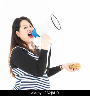 A young fat woman is talking to a megaphone and there is a hamburger in her hand. She is smiling and happy to eat the food she likes. She is hungry. H Stock Photo