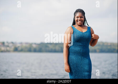 African american dark skinned plus size model posed in a blue shiny dress  against sea side Stock Photo - Alamy