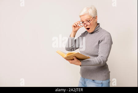 Shocked senior woman reading book and taking off glasses in amazement Stock Photo