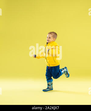 A full length portrait of a bright fashionable boy in a raincoat running and having fun on yellow studio background. Autumn and spring fashion for kids. Cute stylish boy in colorful clothes. Stock Photo