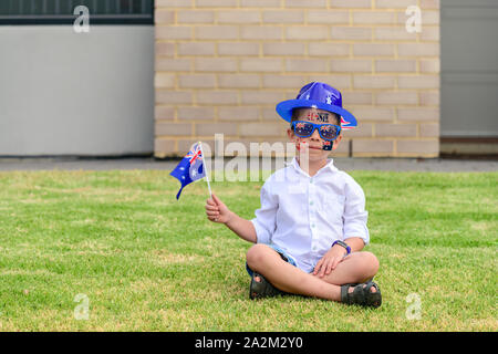 Australian boy wearing hat and sunglasses sitting on green lawn during Australia Day celebration Stock Photo