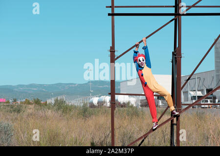 a scary clown wearing a colorful yellow, red and blue costume outdoors, hanging from the rusty structure of an abandoned billboard Stock Photo