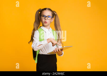 Confused little schoolgirl doing homework on digital tablet, yellow background Stock Photo