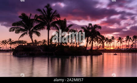 Sun setting on an Hawaiian beach. The sky is purple, red and orange, as the sun dips behind the silhouetted palm trees, on Oahu, Hawaii Stock Photo