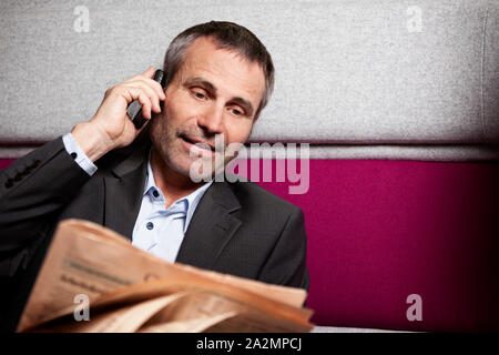 Businessman chatting on mobile phone and reading newspaper. Stock Photo