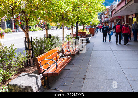 Wooden benches and trees lining the sidewalk with people walking along the shops on Banff Avenue in fall, Banff, Alberta, Canada Stock Photo