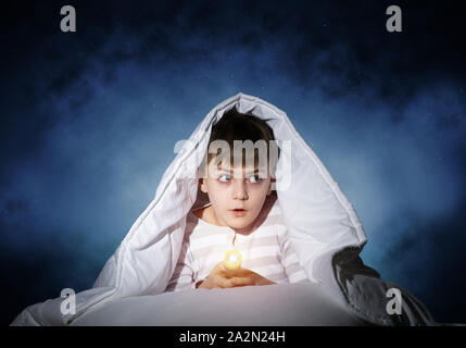 Surprised child with flashlight hiding under blanket. Amazed kid lying in bed at home. Fear of the dark. Little boy can not sleep at night. Portrait o Stock Photo