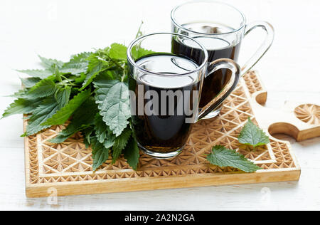 Nettle tea or decoction in glass cups, fresh herbal leaves on wooden cutting board nearby, Urtica dioica is a polyvitaminic dietary food and a cosmeti Stock Photo