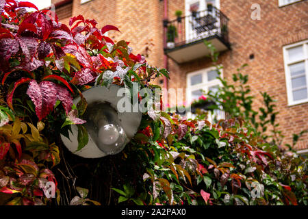 A lamp overgrown with autumn leaves in red, yellow and green colors next to a red brick building. Stock Photo